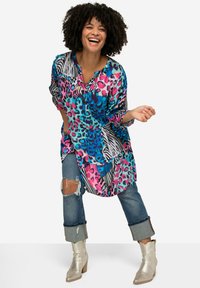 Camisa colorida con estampados de animales en azul, rosa y negro; corte holgado; mangas largas; combinada con jeans desgastados y botas de tobillo plateadas brillantes.