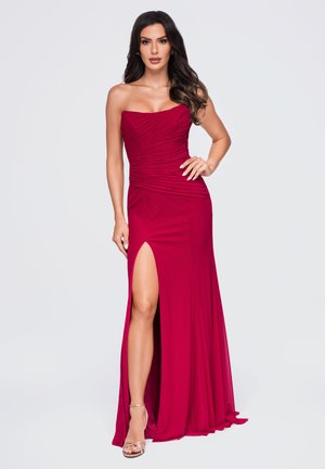 Vrouw met lang donker haar die een strapless rode avondjurk draagt met een split tot aan de dij en zilveren sandalen met hoge hakken, staand tegen een witte achtergrond.