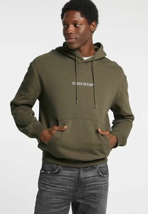 Uomo che indossa una felpa con cappuccio verde oliva con il logo "GUESS JEANS", mani nella tasca frontale, abbinata a jeans neri sbiaditi, guarda di lato.