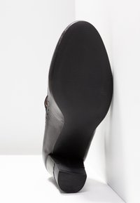 Vue de la semelle d'une chaussure de ville noire, dessous lisse avec un petit talon empilé, positionnée debout contre un coin blanc en arrière-plan.