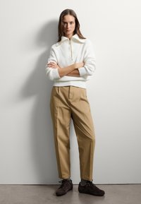 Pull en tricot crème avec un col zippé, associé à un pantalon tapered beige clair. Des chaussures à lacets marron complètent la tenue.