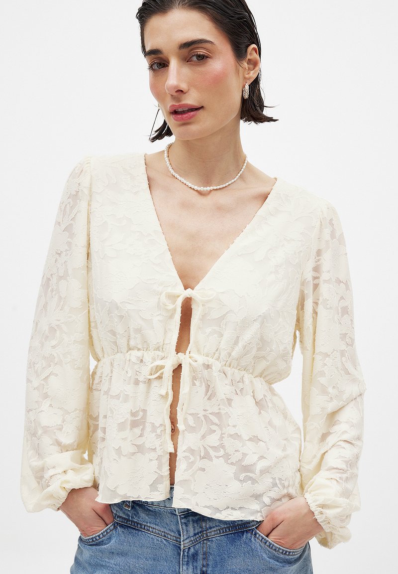 Langärmlige, cremefarbene Bluse aus blumiger Spitze mit tiefem V-Ausschnitt, vorderem Bindedetail und elastischer Taille, kombiniert mit hellblauen Jeans.