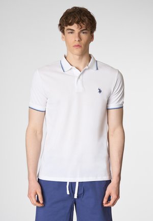 Giovane uomo con capelli ricci indossa una polo bianca con accenti blu e un piccolo logo, sullo sfondo di una superficie chiara.