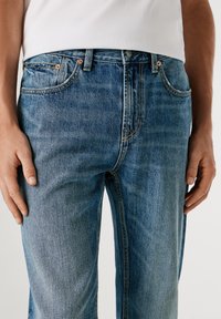 Heledad sinine teksapüksid sirge sääre disainiga, millel on vasknõelad, kontrastne kollane õmblus ja viie tasku stiil.