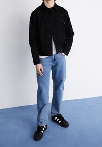 Veste noire boutonnée, jean bleu et baskets noires avec des bandes blanches. La veste comporte une poche poitrine et un col.