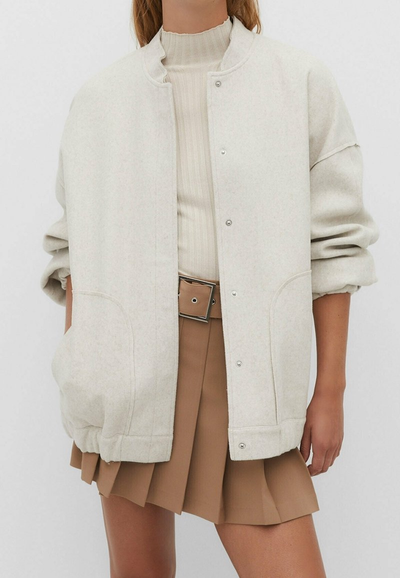 Lichtgrijze oversized jas met een ronde kraag, drukknoopsluiting en zijzakken, gecombineerd met een ribgebreide beige top en een bruine plissé rok.