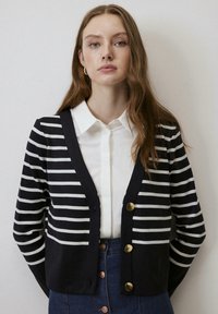 Navy gestreepte cardigan met witte strepen, met een v-hals en drie gouden knopen. gedragen over een witte overhemd met kraag.