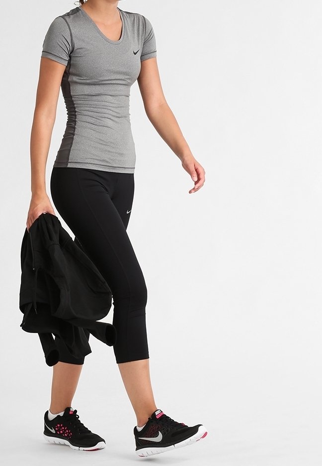 Mujer caminando con leggings cortos negros, camiseta ajustada gris de manga corta, zapatillas negras, sosteniendo una chaqueta negra con una mano.