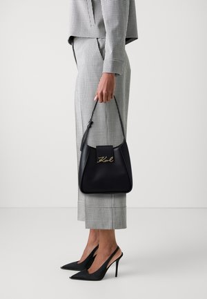 Sac à main en cuir noir avec un logo en script doré, de forme carrée, à anse supérieure, associé à une tenue à carreaux noir et blanc et des talons pointus.