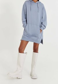 Robe sweat à capuche bleu clair oversize avec poche avant, fentes latérales et capuche à cordon ; associée à des bottes blanches à plateforme montant jusqu'aux genoux.