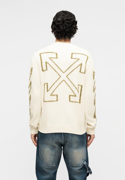 Άνδρας που φοράει λευκό πουλόβερ με μεγάλα υφασμάτινα χρυσά βέλη που σχηματίζουν ένα Χ στην πλάτη, συνδυασμένο με ξεβαμμένα μπλε τζιν.