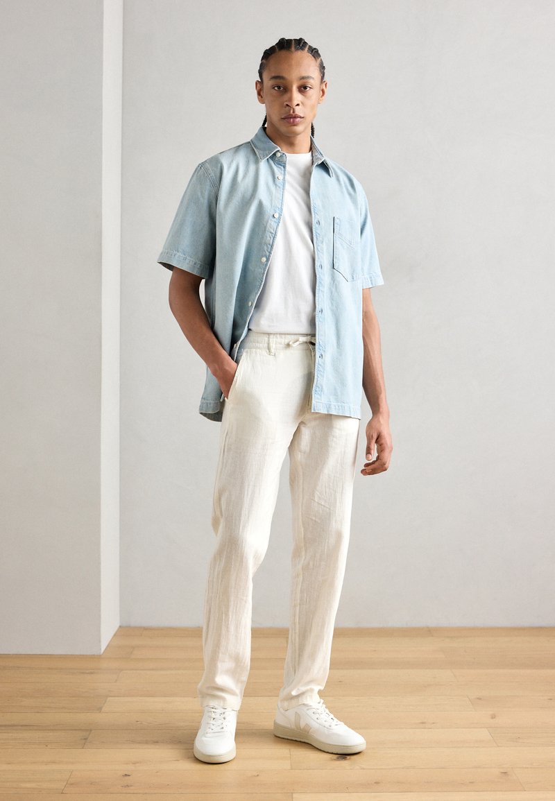Camisa de botones de manga corta azul claro, superpuesta a una camiseta blanca, combinada con pantalones de lino blanco texturizados y zapatillas blancas.