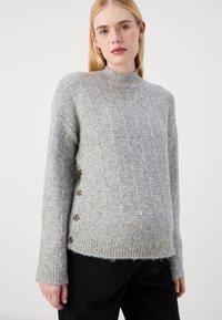 Golf grigio con texture, collo alto, bottoni laterali e polsini a coste, realizzato in un tessuto a maglia. Il fondo è leggermente sfilacciato.