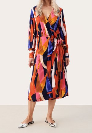 Buntes Wickelkleid in Marineblau, Pink, Orange und Weiß mit geometrischen Mustern. Mit langen Ärmeln und Taillengürtel. Kombiniert mit silbernen Slingback-Schuhen.