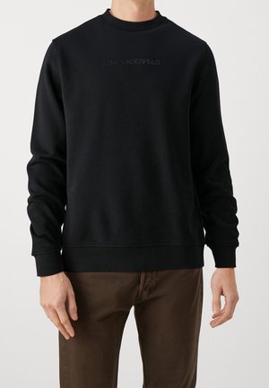 Mann trägt einen schwarzen Langarm-Sweatshirt mit dem Text "Karl Lagerfeld" in kleinen Nieten quer über der Brust und braune Hose.