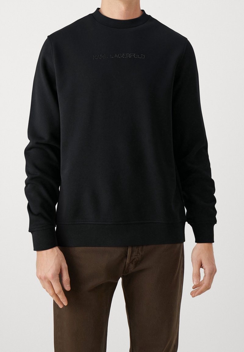Homme portant un sweat-shirt noir à manches longues avec le texte "Karl Lagerfeld" en petites clous sur la poitrine et un pantalon marron.