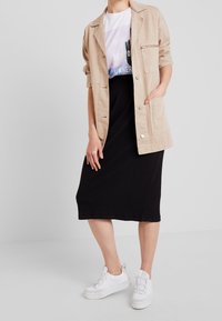 Veste oversized beige, t-shirt graphique blanc, jupe midi noire côtelée et baskets blanches. Tissu lisse avec des coutures visibles et des poches.