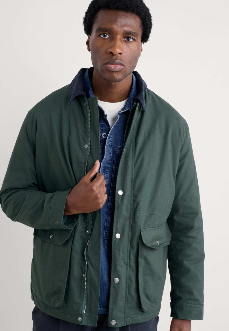 Chaqueta de algodón verde con cierre de cremallera y a presión, con dos bolsillos frontales y un cuello azul, superpuesta a una camisa de mezclilla azul.