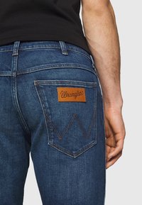 Mörkblå denimjeans med rak passform, som har en brun läderlapp med "Wrangler"-logotyp och ett karakteristiskt sömmönster på bakfickorna.
