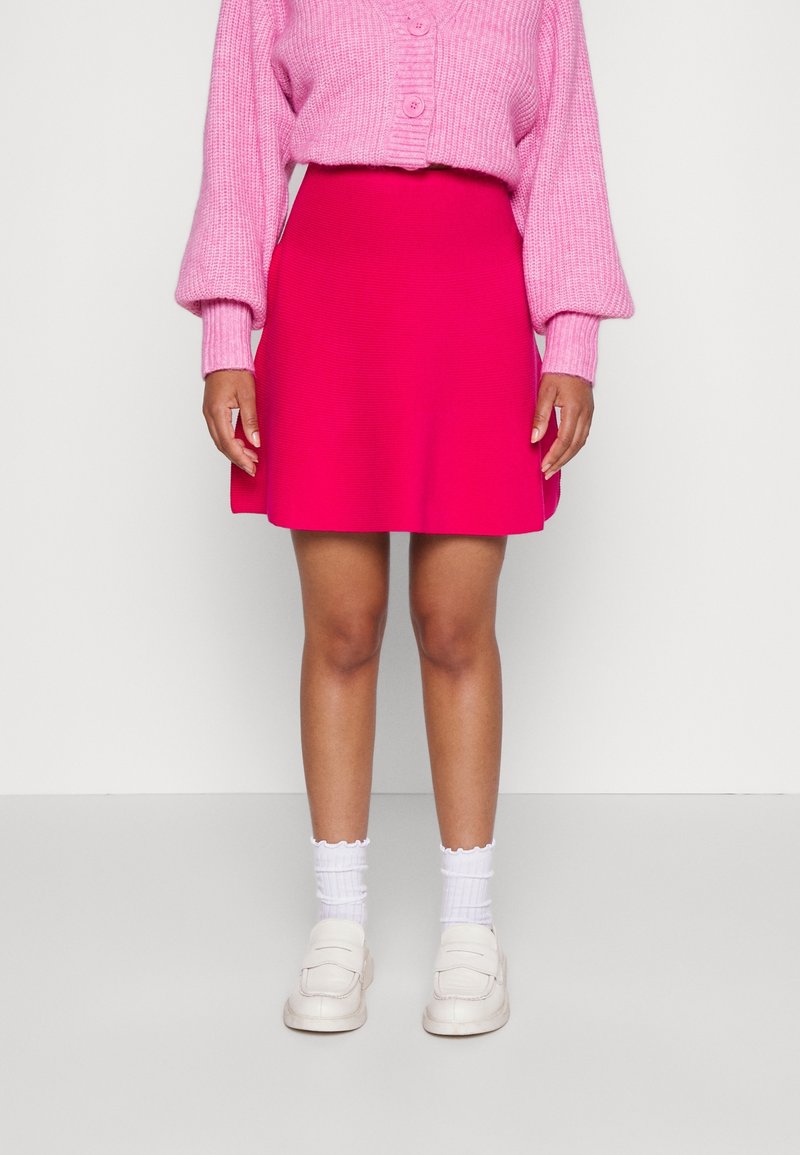 Falda ajustada de color rosa brillante con textura suave. Combinada con un cárdigan de punto rosa holgado y calcetines blancos, usados con zapatos de color claro.