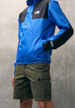Personne portant une veste North Face bleu et noir, les mains dans les poches, associée à un short cargo vert olive devant un mur uni.
