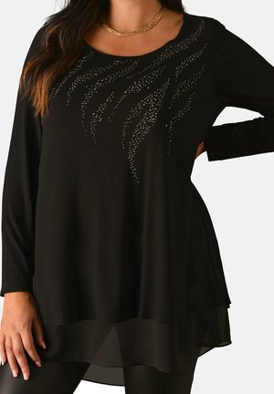 Schwarze Tunika mit langen Ärmeln und silbernem Strass-Wellenmuster auf der Brust, transparentem Saum, getragen von einer Person mit Hand in der Hüfte und einer goldenen Halskette.