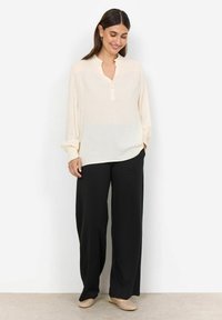 Light beige long-sleeve blouse with a mandarin collar, button front, and gathered cuffs, paired with black wide-leg trousers and woven slip-ons.