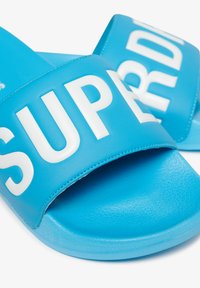 Blauwe slidesandalen van glad synthetisch materiaal met een brede band waarop de tekst "SUPR" in grote witte letters staat. Geprofileerde zool.