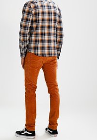 Rutigt skjorta i marinblått, orange och vitt; bruna kordfloretbyxor; svarta sneakers med vita accenter; sedd bakifrån.