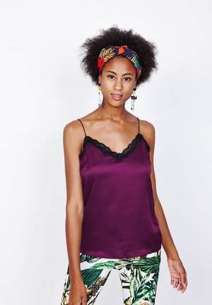 Mujer joven con peinado afro que lleva una diadema colorida, camisola morada con encaje y pantalones blancos con estampado de hojas verdes, posando.