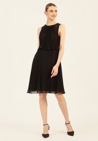 Schwarzes plissiertes Kleid mit ärmellosem Design, das einen geschichteten Oberteil und einen halb durchsichtigen Saum aufweist. Getragen mit schwarzen spitzen Absätzen.