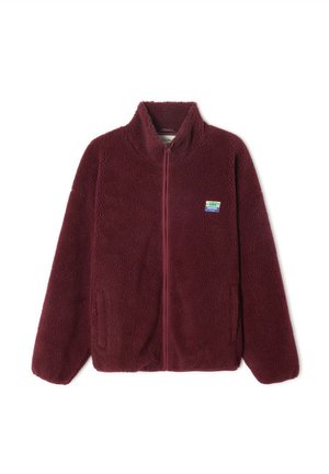 Veste polaire bordeaux avec un col montant, une fermeture éclair à l'avant, des poches latérales et un petit patch coloré sur la poitrine gauche. Texture douce.