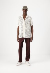 Chemise à manches courtes avec un motif en dentelle de couleur crème, associée à un pantalon taillé bordeaux et des baskets blanches. Une casquette en vert discret vient compléter le look.