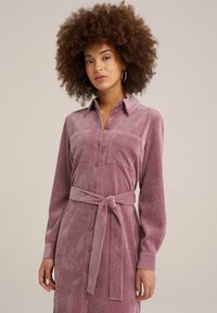 Rosa kordflodskjol med lång ärm, knappstängning fram, två bröstfickor och ett knytbälte i midjan för en figurnära siluett.