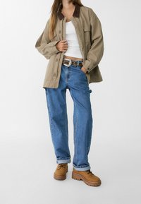 Beige Jacke mit braunem Kragen, weißem Tanktop, blauen weit geschnittenen Jeans und braunen Schnür-Plateauschuhen. Der Gürtel hat dekorative Akzente.