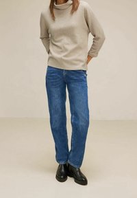 Beige randig tröja med halskrage, avslappnad passform, ihopparad med blå rakbeniga jeans och svarta anklar stövlar, mot en neutral bakgrund.