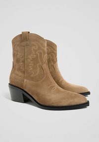 Brązowe zamszowe kowbojki z wąskimi noskami, kontrastującymi białymi szwami i stosowaną czarną obcasem. Zawierają pętle do wciągania na górze.