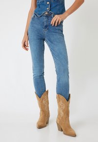 Botas de vaquero de ante marrón con puntas afiladas y costuras decorativas, combinadas con jeans ajustados de mezclilla azul. Las botas tienen un tacón apilado.