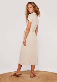 Beige Leinen-Midikleid mit kurzen Ärmeln, einem kragenähnlichen Ausschnitt und einem taillierten Gürtel. Ausgestattet mit einer subtilen Textur und getragen mit braunen Riemchensandalen.