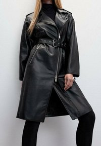 Femme portant un trench-coat en cuir noir ceinturé avec des détails à fermeture éclair, sur des leggings noirs, posant devant un fond clair uni.