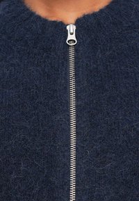 Artimas tamsiai mėlynos vilnonės užtrauktuko striukės vaizdas, su sidabriniu metaliniu užtrauktuku, dalinai užtrauktu šalia kaklo.