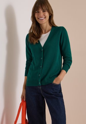 Femme souriante portant un cardigan vert boutonné, une chemise blanche, un jean bleu foncé, tenant une sangle de sac orange avec une main dans sa poche.