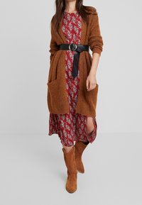 Brun stickad cardigan med fickor över en röd blommig klänning, åtdragen i midjan med ett svart bälte. Ett par tanfärgade ankelboots fulländar looken.