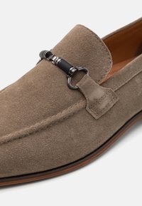 Brown suede loafer with a rounded toe, featuring a black decorative buckle and a tan leather interior. Simple stitched detailing along the edges.