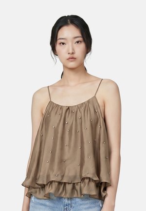 Giovane donna con capelli scuri indossa un top marrone ampio senza maniche con piccole perline decorative e jeans azzurri chiari.