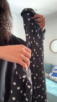 Schwarzes Kleid mit weißen, unregelmäßigen Polka-Punkten, aus einem glatten Stoff. Lange Ärmel, die auf Schulterhöhe gehalten werden, mit einem fließenden Design.