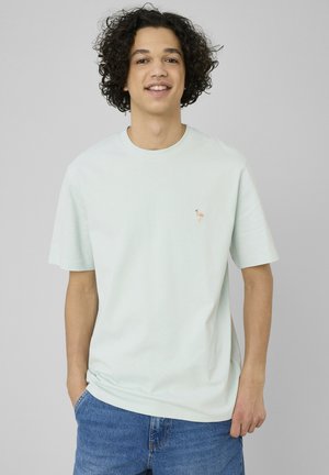 Jeune homme aux cheveux bouclés porte un t-shirt clair avec un petit logo flamant rose, associé à un jean bleu, debout devant un fond uni.
