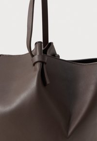 Borsa tote in pelle marrone scuro con sottili manici e un dettaglio di nodo vicino all'attacco dei manici, su uno sfondo chiaro e semplice.