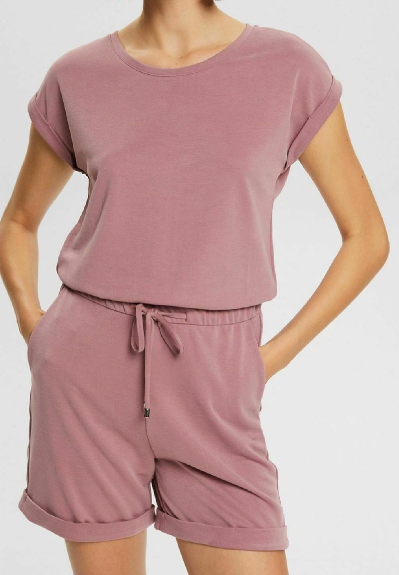 Femme portant un haut à manches courtes mauve assorti et un short à cordon et ourlets retroussés, mains dans les poches, sur fond uni.