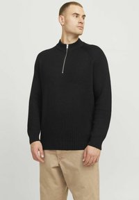 Schwarzer Rippstrick-Pullover mit hohem Kragen und halblanger Reißverschluss. Verfügt über lange Ärmel und eine lockere Passform. Getragen mit beigen Hosen.